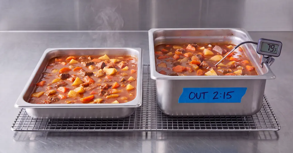 Two hotel pans of stew cooling side by side, a shallow pan and a deeper pan, with a clipped digital thermometer reading 79°F on the deeper pan and a handwritten "OUT 2:15" time label, illustrating how pan depth affects cooling speed.