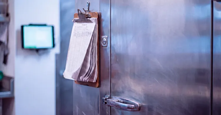 A close-up view of a metal walk-in cooler door with a paper clipboard hanging from the handle and a small digital temperature display mounted on the adjacent wall.