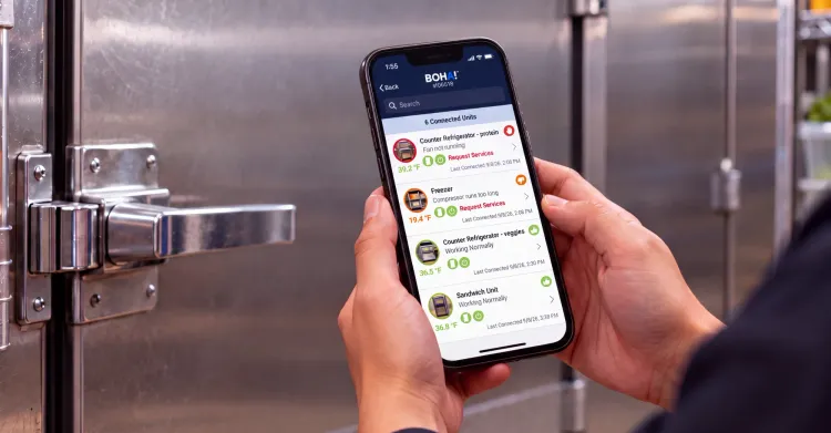 Hands holding a phone showing the BOHA! app with temperature status for connected kitchen equipment, including a refrigerator flagged because its fan isn’t running and a freezer flagged for a long compressor run, in front of a walk-in cooler door.