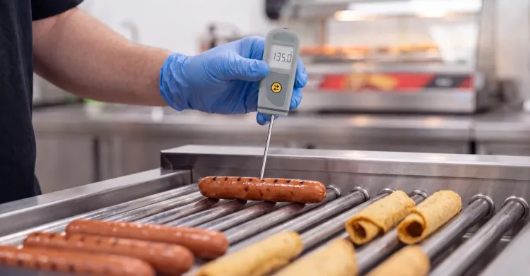 A c-store employee taking a digital thermometer reading of hotdogs. 