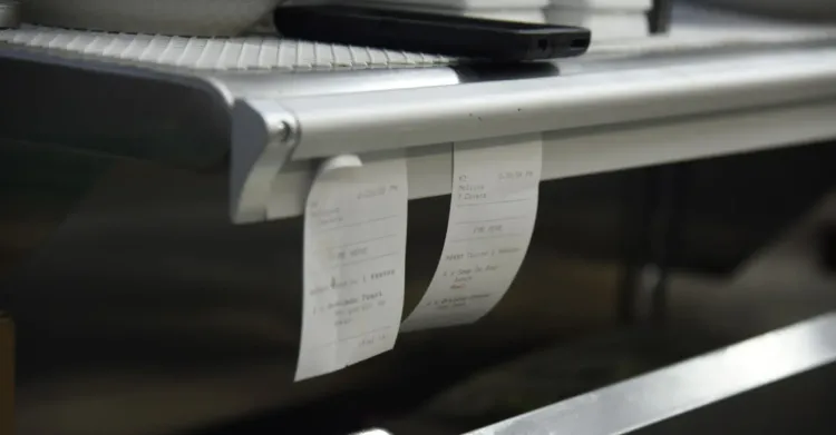Paper tickets hanging in a restaurant back-of-house area, showing manual foodservice workflows that can become harder to manage under operational pressure.