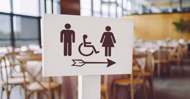 wooden directional sign displaying dark brown male, accessible, and female restroom icons with a right-pointing arrow, set against a blurred background of restaurant seating.