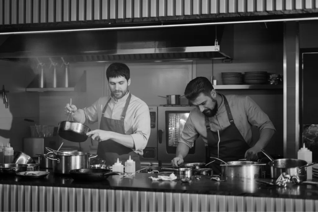 Two chefs working a busy fine-dining kitchen line, in black and white