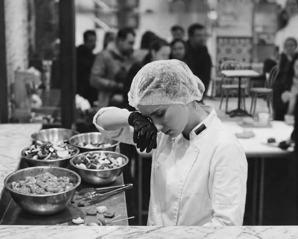 A kitchen worker in a hairnet preparing ingredients at a marble prep counter, in black and white