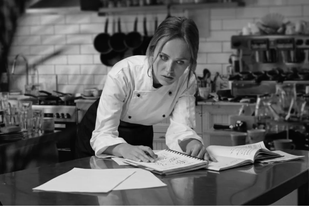 A chef writing in a notebook at a kitchen prep counter, in black and white