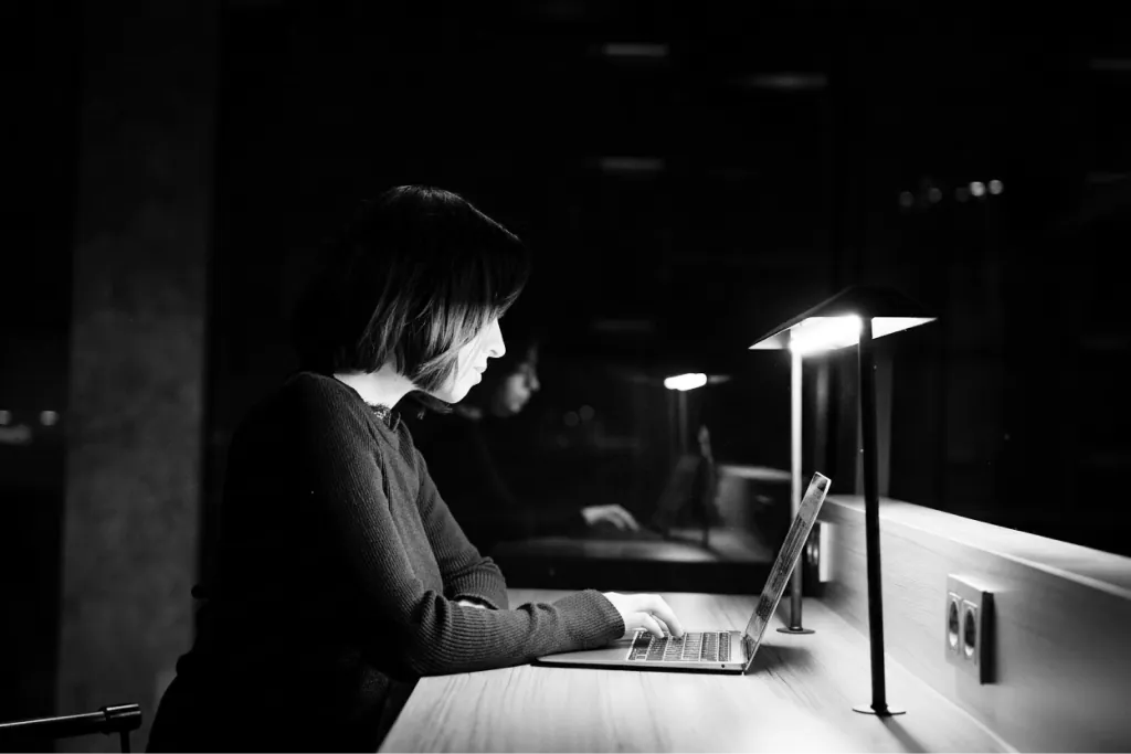 A corporate employee working late alone at a laptop in a dark office, in black and white