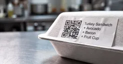 Close-up of a printed made-to-order label with barcode applied to a wrapped sandwich, kitchen blurred in the background
