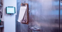 A close-up view of a metal walk-in cooler door with a paper clipboard hanging from the handle and a small digital temperature display mounted on the adjacent wall.