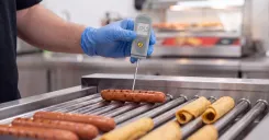 A c-store employee taking a digital thermometer reading of hotdogs. 