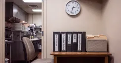 An empty manager's chair beside a desk holding labeled compliance binders, HACCP Plan, Corrective Actions, Training, SDS, and Inspection Reports, with a wall clock above and a commercial kitchen visible through the doorway.