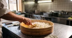 Sushi chef mixing seasoned vinegar into warm sushi rice in a traditional wooden hangiri bowl using a rice paddle.